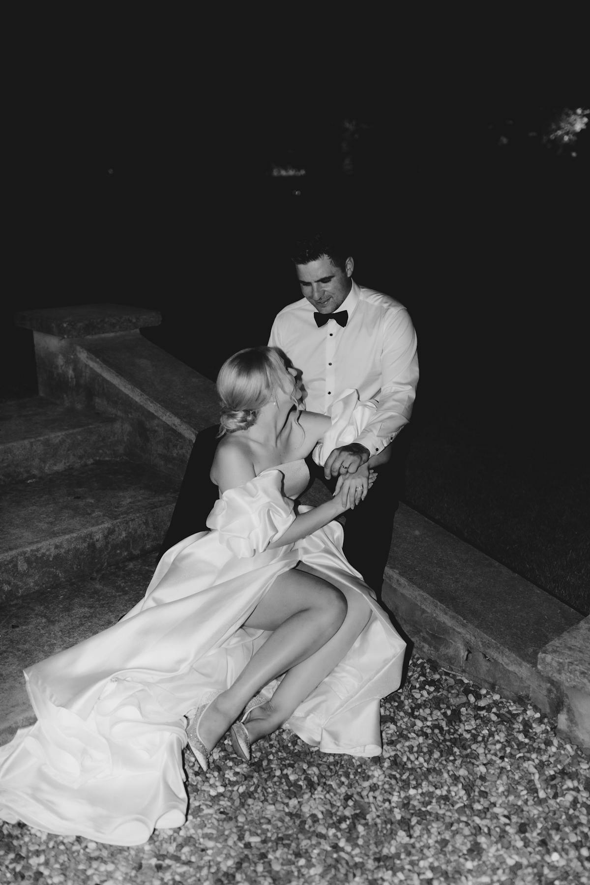 A black and white photo of a couple seated on outdoor steps at night. The bride, in a flowing off-shoulder gown, sits on the groom’s lap and looks at him affectionately. The groom, dressed in formal attire with a bow tie, gazes back at her with a smile.
