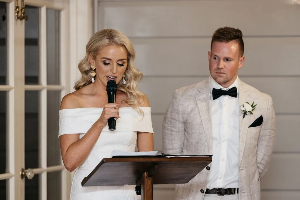 A woman in a white off-the-shoulder dress is speaking into a microphone at a podium. A man in a light-colored suit with a black bow tie stands beside her, looking on attentively. Both appear to be indoors, against a backdrop of light-colored paneling and a glass door.
