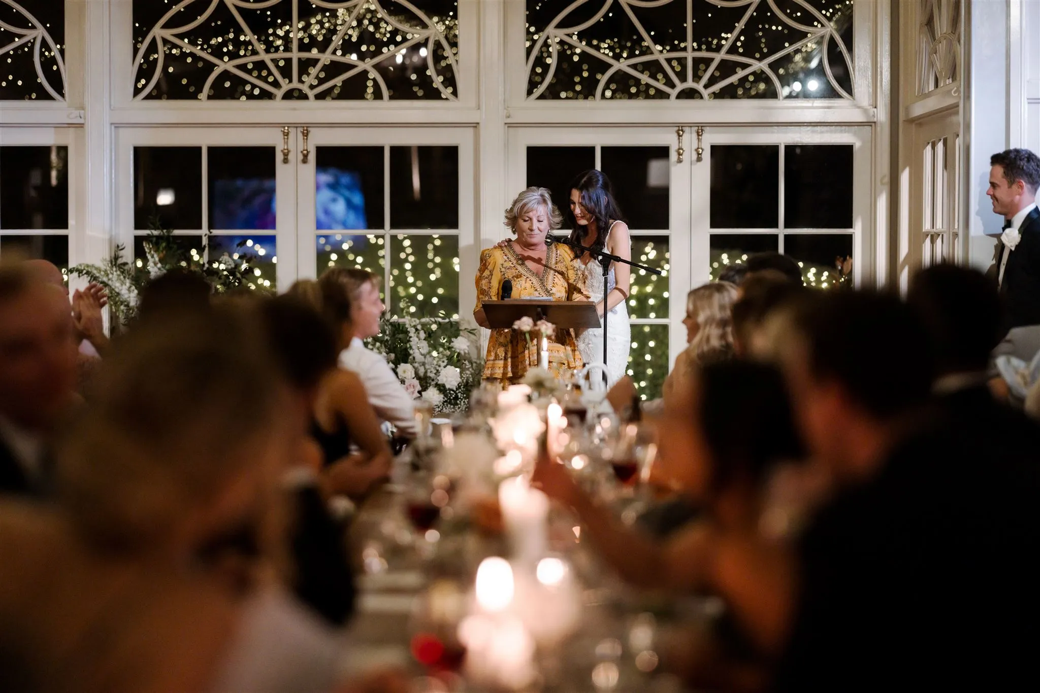 A woman in a gold and black dress stands at a podium delivering a speech, while a second woman in a white dress stands beside her. Guests are seated at dimly lit tables in a decorated venue with fairy lights and arched windows in the background.