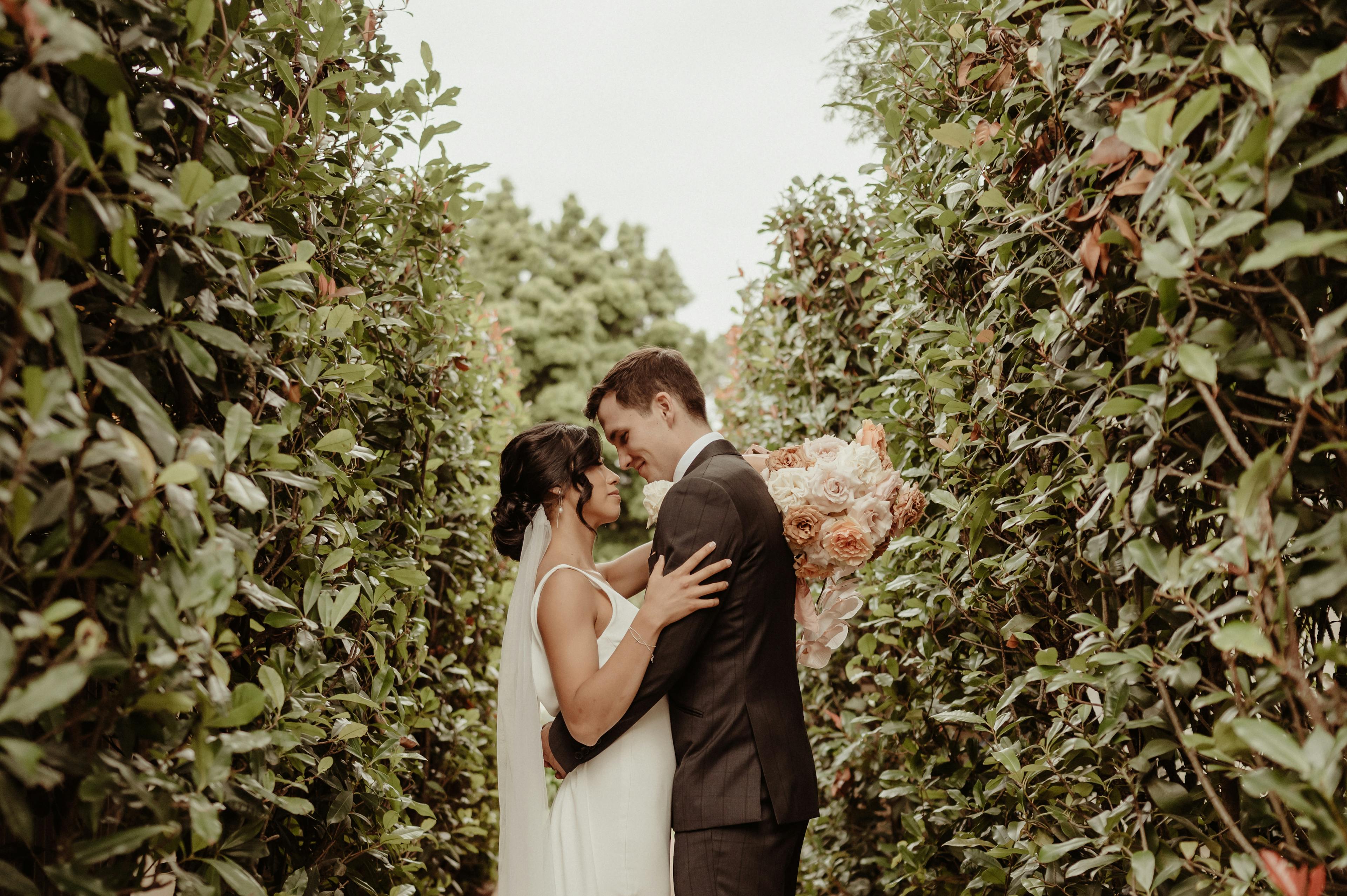 A bride and groom are standing closely in an outdoor setting surrounded by lush greenery. The bride holds a bouquet of peach-colored flowers, and both are gazing lovingly at each other while lightly embracing. The setting appears intimate and serene.
