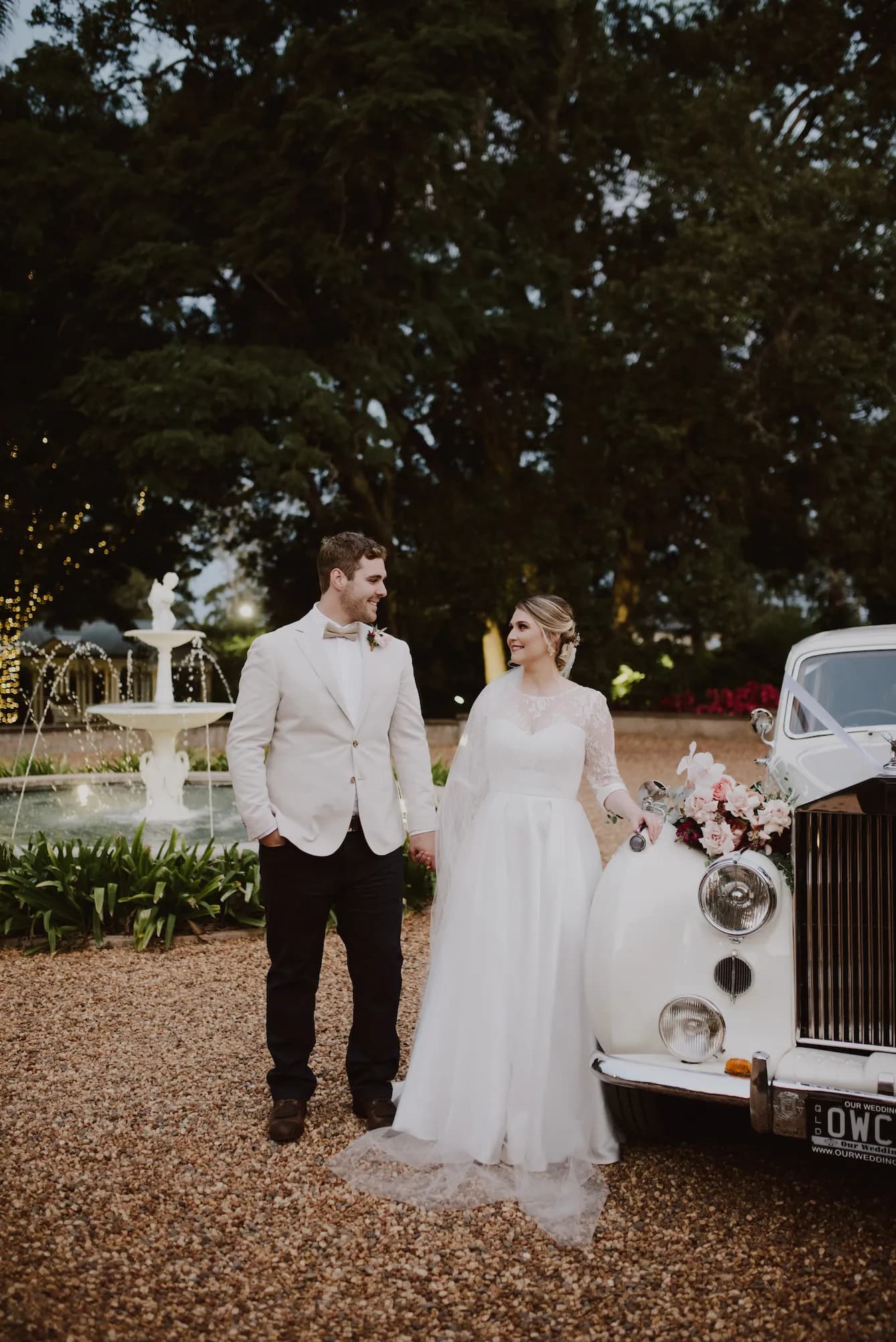 A bride and groom stand next to a vintage white car adorned with flowers. The bride wears a white gown with lace sleeves and a veil, while the groom is dressed in a white jacket and dark trousers. They are smiling at each other with a fountain and large trees in the background.