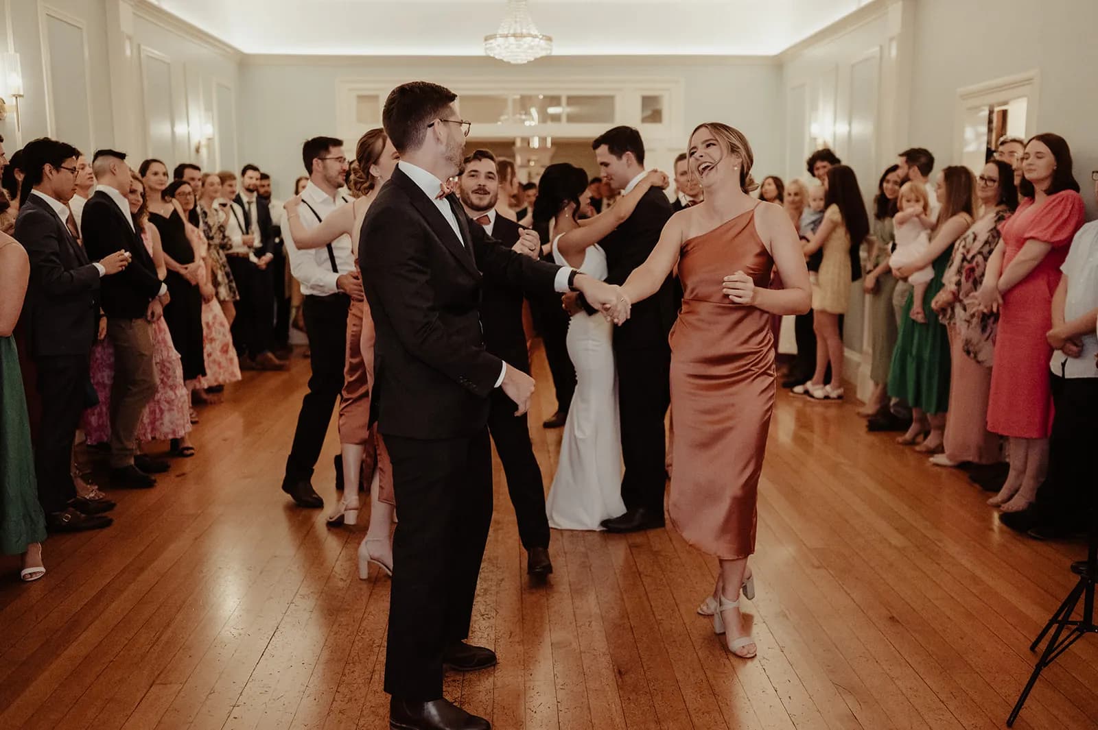 A well-dressed couple dances in the center of a large, elegant room with wooden floors, surrounded by a group of people who are either dancing or observing. The atmosphere appears festive, with chandelier lighting adding to the formal ambiance.
