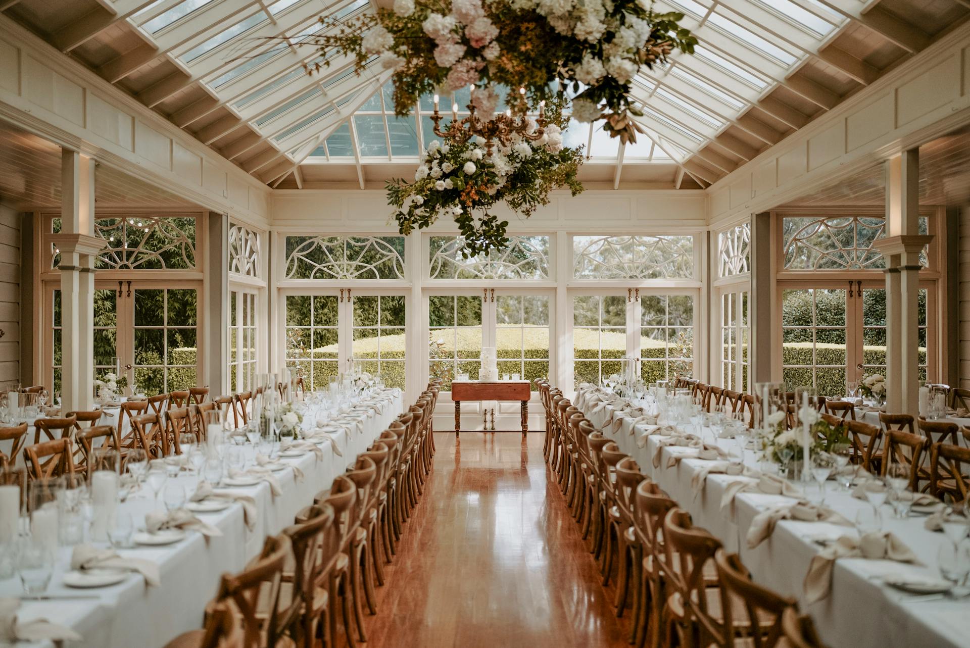 A beautifully decorated event space with a glass ceiling and large windows, featuring two long banquet tables set with white linens, glasses, and floral centerpieces. Wooden cross-back chairs line the tables, and a chandelier adorned with flowers hangs from above.