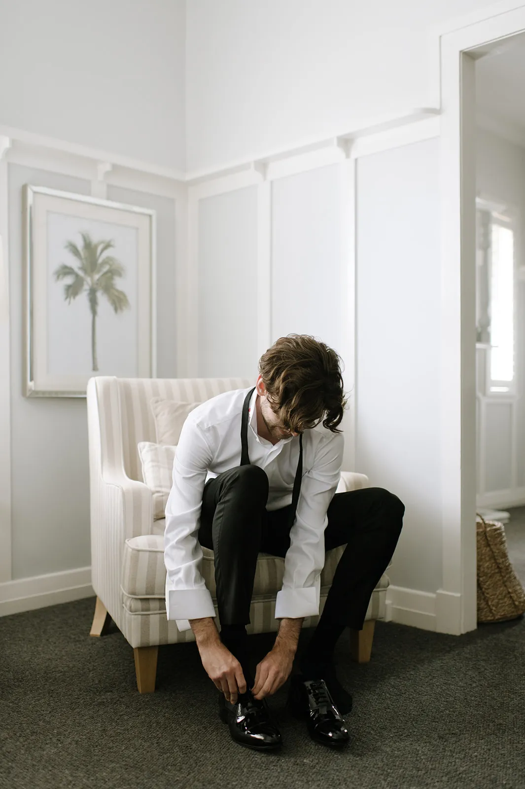 A man in a white dress shirt and black pants, with a black bow tie untied around his neck, sits on a beige-striped armchair. He is bending down, tying his shiny black shoes. The room has minimalist decor with a palm tree painting on the wall.