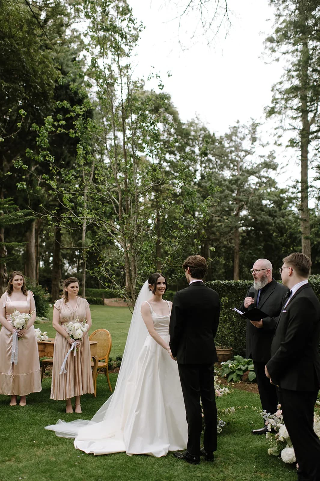 A couple stands facing each other at an outdoor wedding ceremony with a lush green garden backdrop. The bride is in a white gown and veil, and the groom is in a black suit. Bridesmaids and groomsmen stand nearby, and a bearded officiant reads from a book.