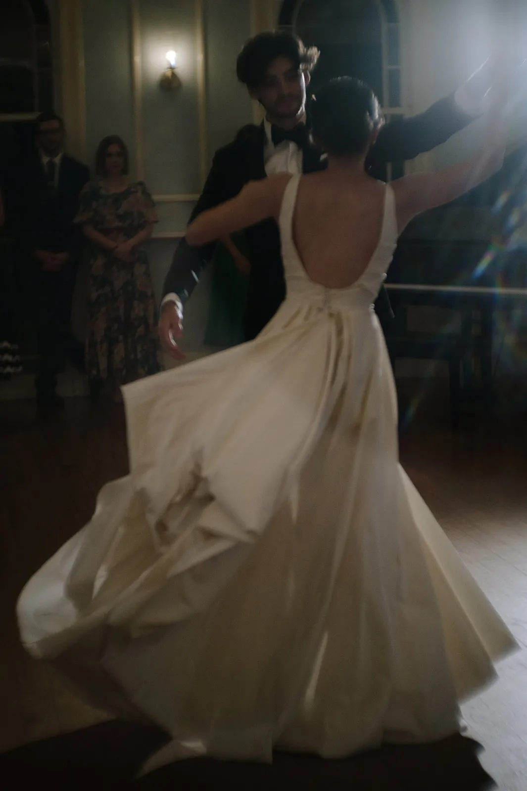 A couple is dancing elegantly in a dimly lit room. The woman is wearing a flowing white gown with a low back, and the man is dressed in a dark suit. Other guests are faintly visible in the background, watching the pair as they twirl across the dance floor.