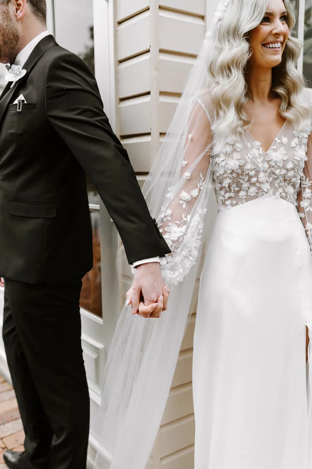 Bride and groom reachng around wall to hold hands