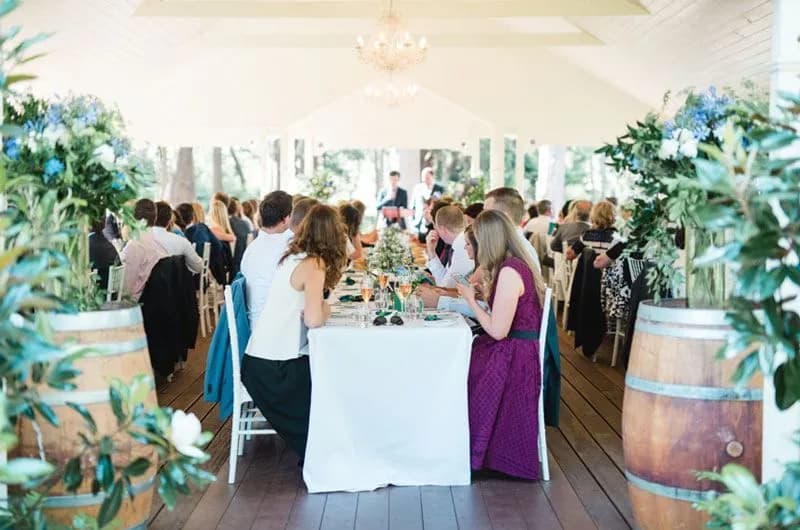 A large gathering of people sits at long tables in a bright, elegant venue with high ceilings. The tables are adorned with white tablecloths and floral arrangements. Wooden barrels and lush greenery decorate the space, adding to the sophisticated atmosphere.