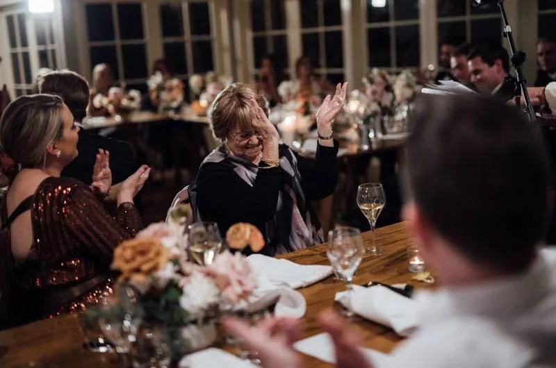 A group of people sit at decorated tables in a warmly lit room. A woman at the center, wearing glasses and a scarf, covers her face with her hand while clapping with the other. Others around her smile and clap, creating a joyful and celebratory atmosphere.