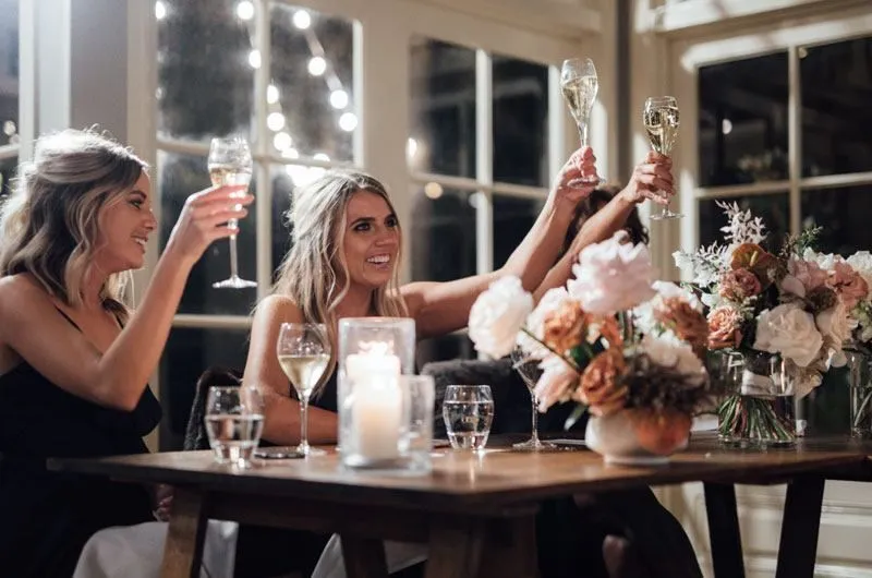 Three women in elegant dresses toast with champagne glasses at a beautifully decorated dinner table adorned with flowers and candles. The scene is warm and celebratory, with soft lighting and a festive atmosphere.