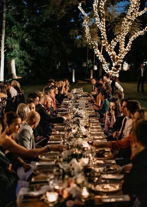 A long outdoor dining table is set for a formal event, surrounded by guests in semi-formal attire. The table is adorned with candles and floral arrangements. String lights are draped around a tree in the background, creating a warm, festive atmosphere.