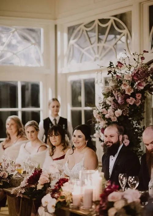 A wedding reception with a smiling bride and groom seated at a long table adorned with floral arrangements and candles. Bridesmaids and a groomsman are seated alongside them. A waiter stands behind them in a room with large windows and elegant decor.