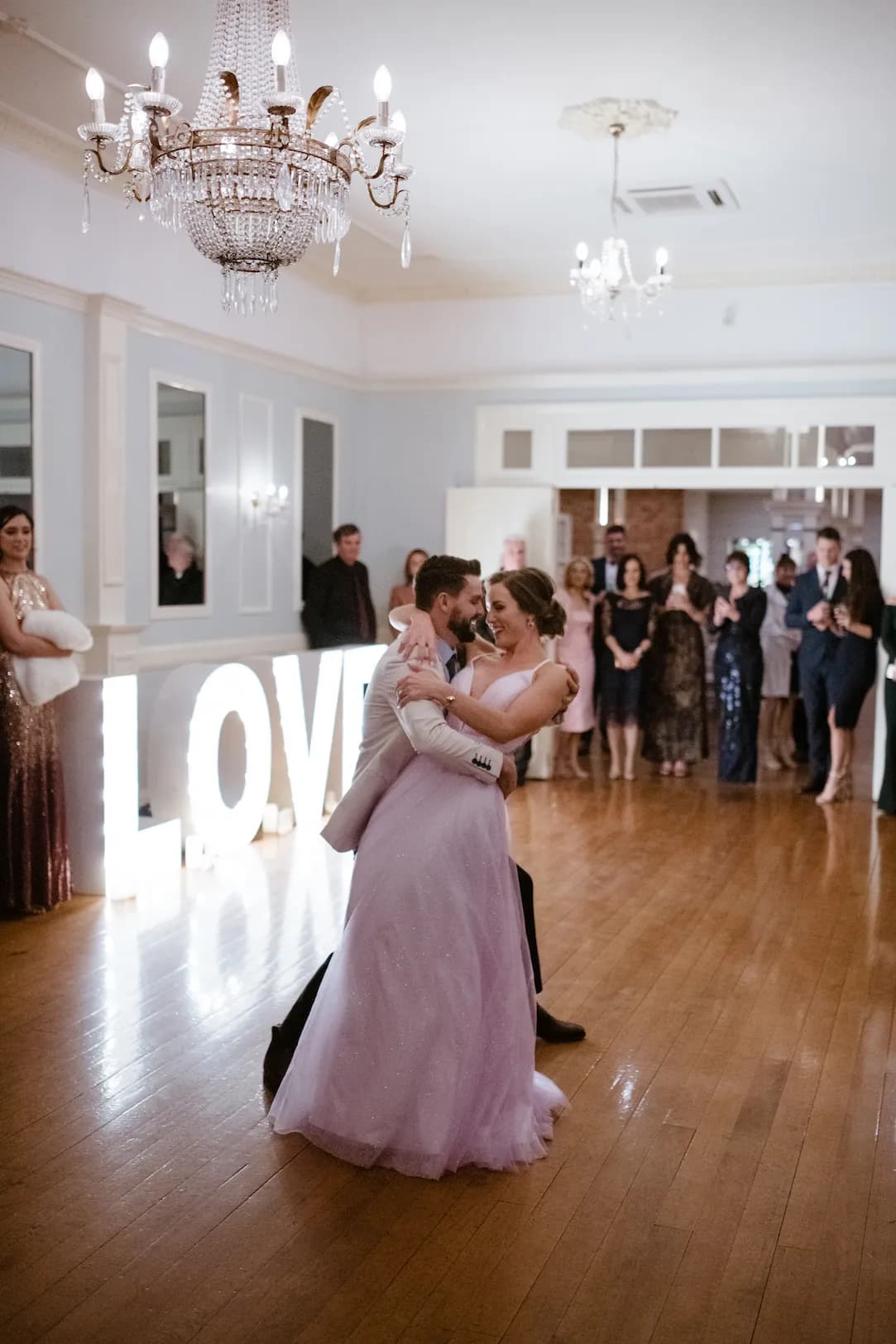 A couple shares a romantic first dance at their wedding reception. The bride in a flowing white gown and the groom in a sharp suit are surrounded by smiling guests in elegant attire. A large illuminated "LOVE" sign and chandeliers add to the elegant ambiance.