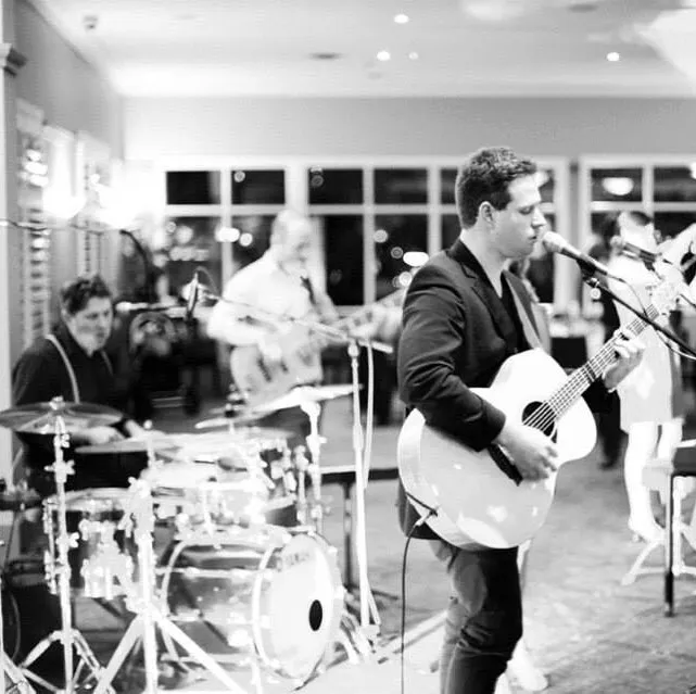 Black and white photo of a small band performing indoors. The focus is on a man in a dark suit singing and playing an acoustic guitar at the mic. A drummer and a guitarist in the background are also visible, with blurred audience members and lights behind them.