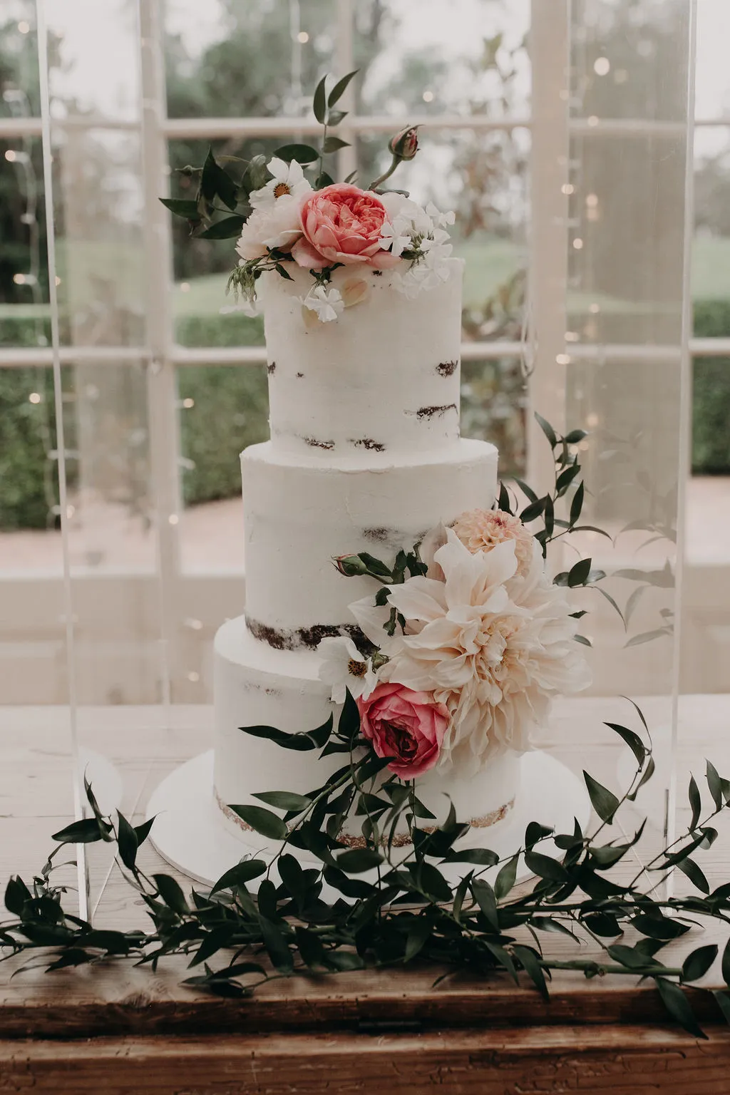 A three-tiered white wedding cake adorned with pink and white peonies, delicate small flowers, and green leaves. The cake is placed on a wooden table with additional greenery around the base. Soft outdoor light filters through a window in the background.