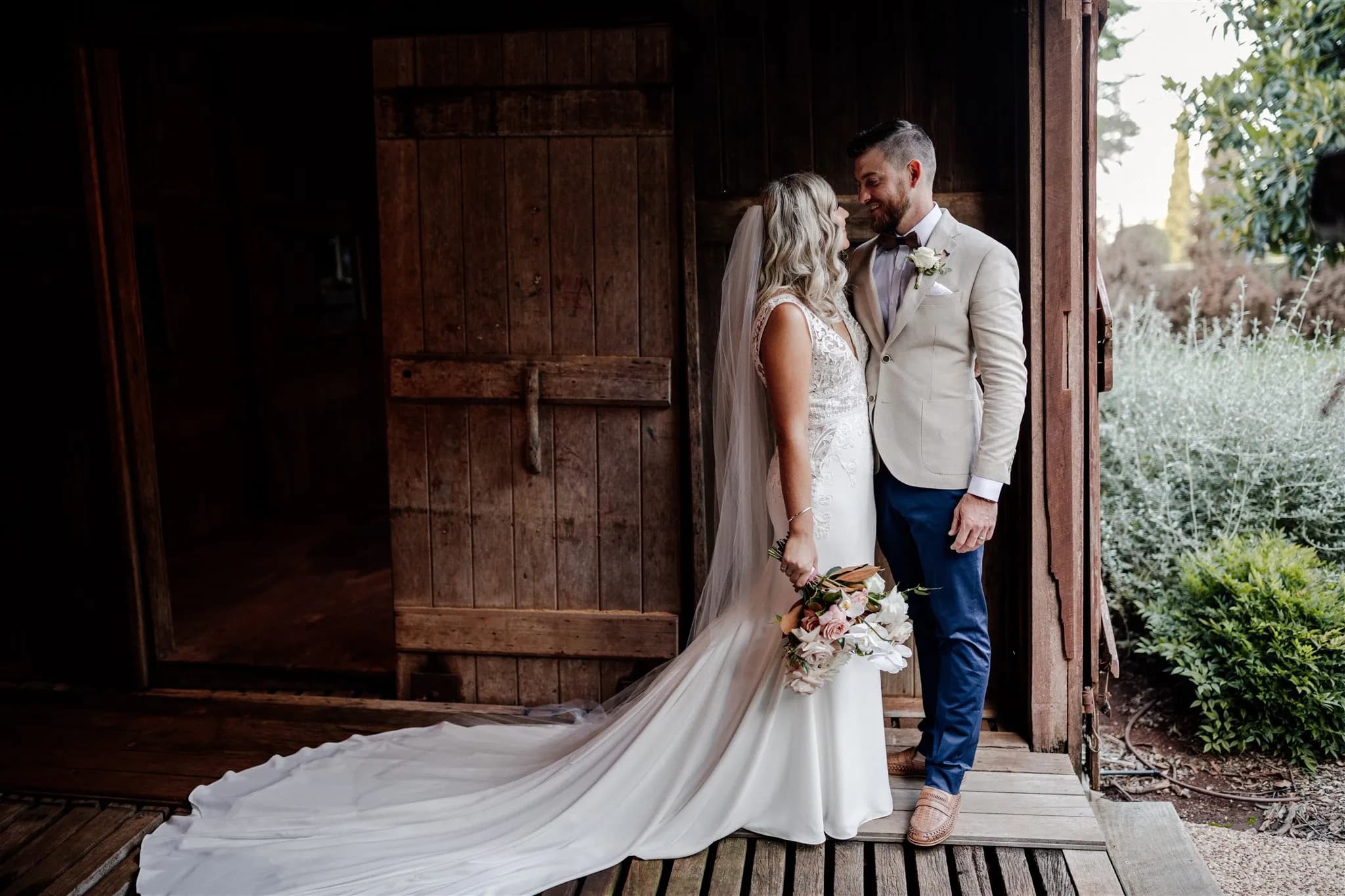 A couple dressed in wedding attire stand close to each other beside a wooden building. The bride, in a long white gown with a train and holding a bouquet, smiles at the groom in a light-colored jacket and dark pants. They appear happy and serene.