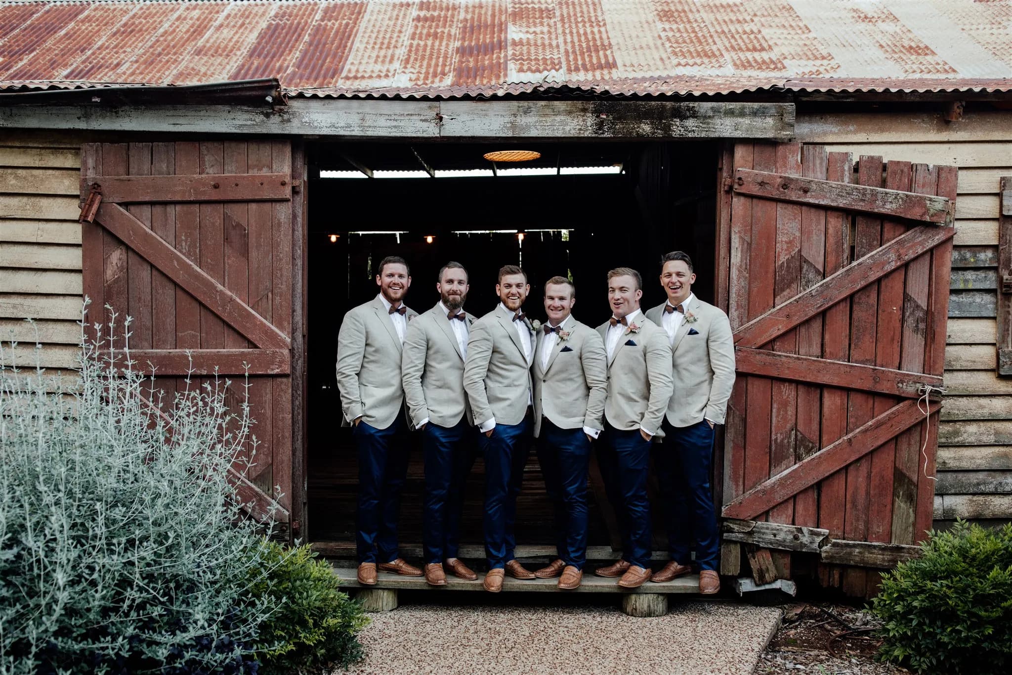 Six men dressed in light-colored jackets, white shirts, dark pants, and brown shoes stand side-by-side in front of a rustic, open barn door. They are smiling and have boutonnières on their jackets. The barn has wooden walls with a corrugated metal roof.