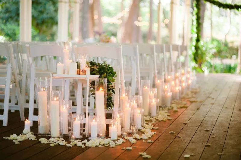 An elegant wedding aisle lined with white chairs, illuminated by numerous lit candles of varying heights in glass holders. White rose petals are scattered along the aisle, and greenery decorates the columns, creating a romantic ambiance.