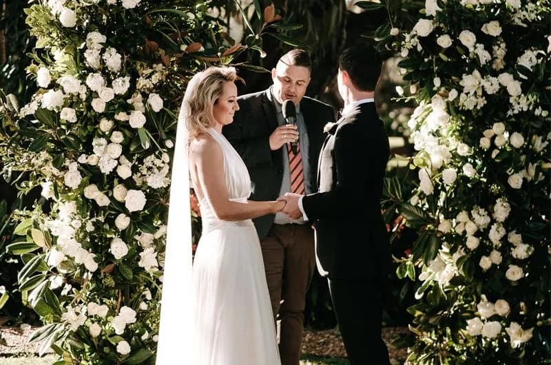 A bride and groom, dressed in formal attire, stand holding hands facing each other under a floral archway adorned with white roses. A suited man stands behind them, speaking into a microphone. The outdoor setting is lush and green.