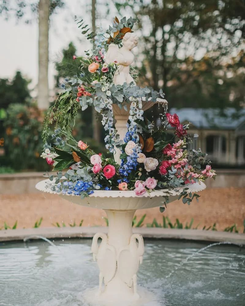 A beautiful white stone fountain with water flowing over the tiers is adorned with vibrant, fresh flowers in pink, blue, and white. Greenery and a small statue of an angel embellish the top, set against a lush garden backdrop.
