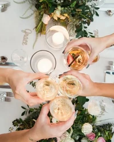 Overhead view of four hands holding glasses in a toast, above a decorated dinner table. The centerpiece features flowers and greenery, with lit candles adding a cozy ambiance. The table is set with cutlery and plates.