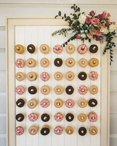 A donut wall displays an assortment of donuts in three different varieties: plain, chocolate, and pink with white icing stripes. The wall is adorned with a bouquet of red, white, and pink flowers in the top right corner.