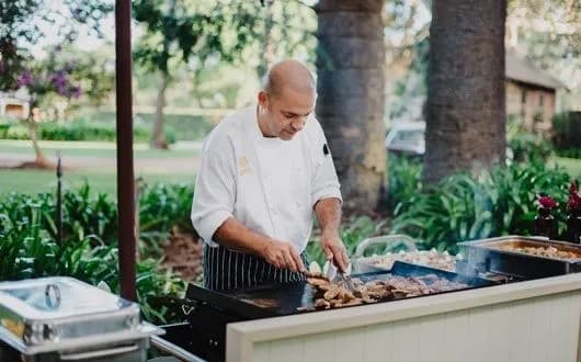 A chef in a white uniform and striped apron is grilling meat on a large outdoor grill. He is surrounded by lush greenery and trees in a garden setting. The chef is focused on turning the meat with tongs.