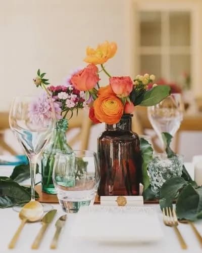 A table setting featuring a centerpiece with vibrant flowers in an amber bottle, surrounded by empty wine glasses, a water glass, and gold utensils on a white tablecloth. The flowers include shades of orange, pink, and yellow, alongside green foliage.