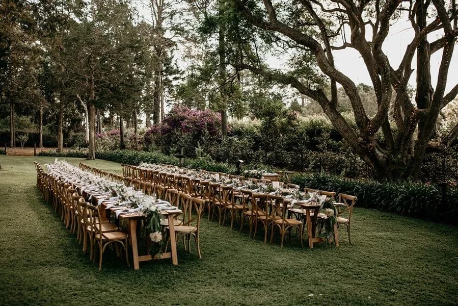 An outdoor wedding reception setup featuring long wooden tables adorned with white tablecloths, floral arrangements, and greenery runners. Wooden chairs are neatly arranged around the tables, all placed on a lush green lawn surrounded by tall trees and flowering bushes.
