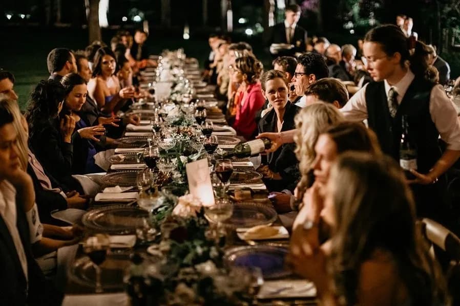 A large group of people is seated at a long, elegantly set dinner table for an outdoor evening event. The table is decorated with flowers and candles. A waiter is pouring wine, and attendees are engaged in conversation, creating a lively and festive atmosphere.