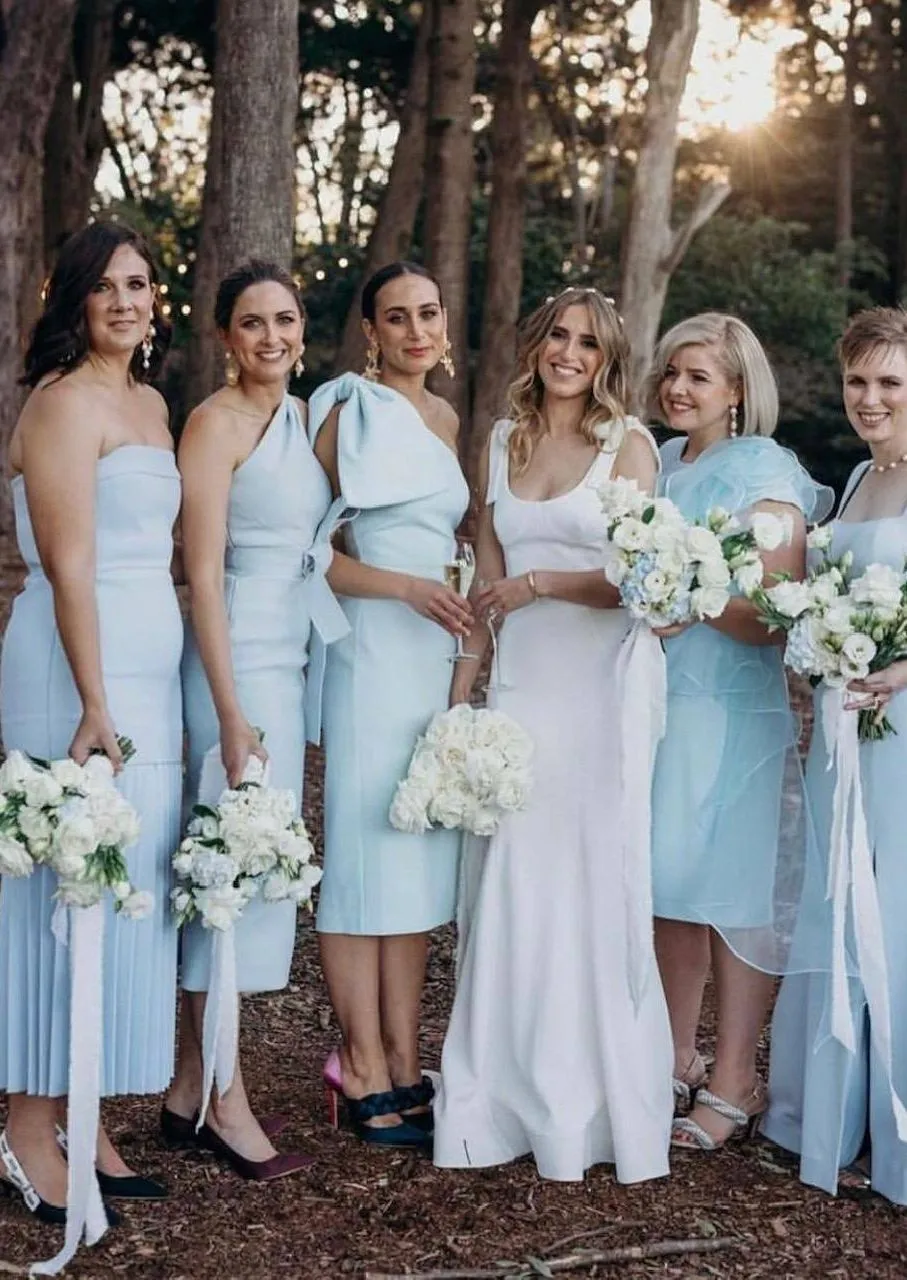 A bride in a white dress stands outdoors with five bridesmaids wearing light blue dresses. They are all holding bouquets of white flowers with greenery. Trees and sunlight can be seen in the background, creating a serene and natural setting.