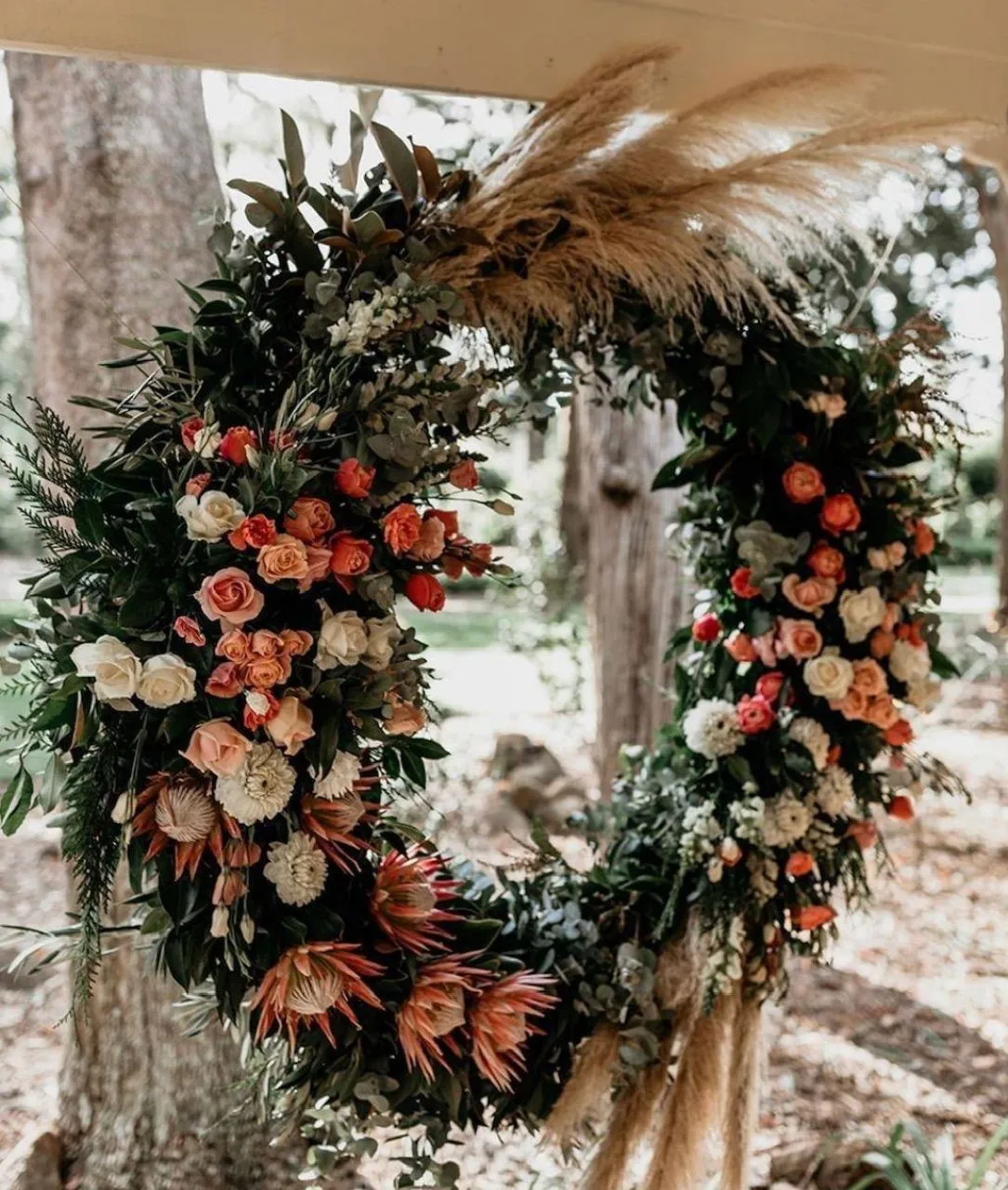 A large circular floral arrangement hangs outdoors. It features a variety of flowers, including roses, proteas, and greenery, accented with wispy pampas grass. The background shows trees and a garden setting, giving a natural, rustic ambiance.