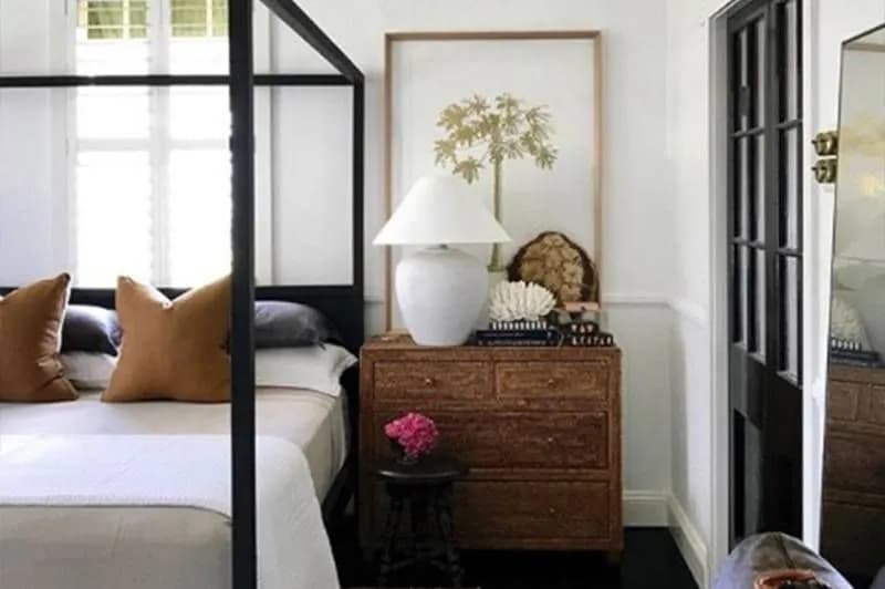 A cozy bedroom features a black four-poster bed with white linens and tan pillows. Beside it, a wooden nightstand holds a white table lamp, books, and a decorative seashell. A framed botanical print hangs above, with a small pink flower on a nearby dark side table.
