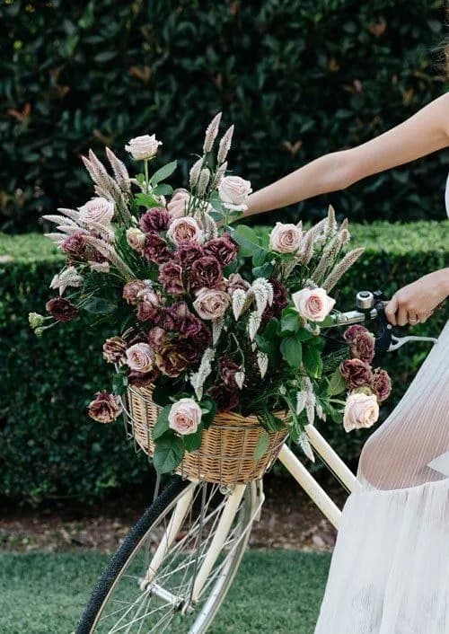A person wearing a white dress is holding a bicycle with a large woven basket on the front. The basket is filled with an arrangement of pink and dark purple roses mixed with various greenery and wheat-like plants. There is a lush garden hedge in the background.