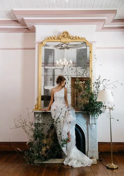 A bride in a white, one-shoulder wedding gown sits on a marble fireplace mantle, adorned with greenery. Above her is a large, gold-framed mirror reflecting a chandelier. The room has light-colored walls, a floor lamp, and wooden floors.
