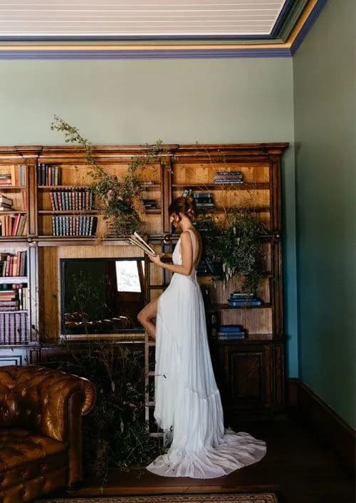 A woman in a flowing white gown stands on a ladder, reaching for a book from a wooden bookshelf filled with books. The bookshelf is adorned with green vines and plants, adding a touch of nature to the elegant and classic room. A leather sofa is partially visible.