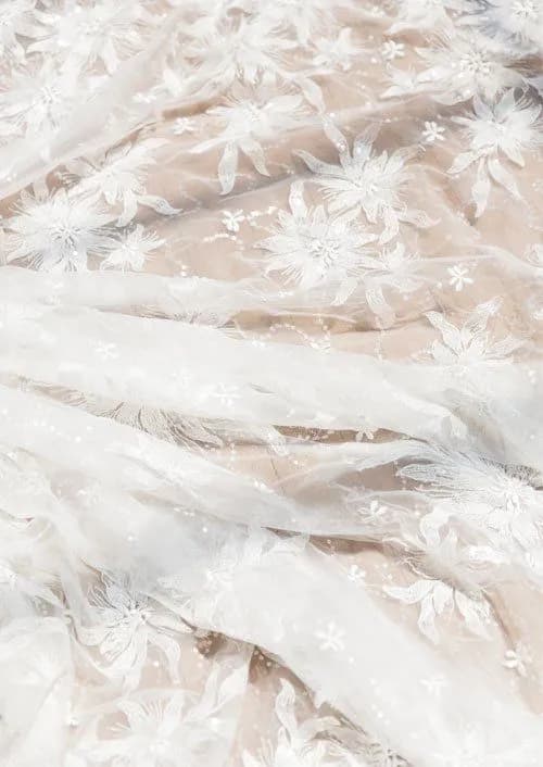 Delicate white floral lace fabric is spread out, showcasing intricate flower patterns and translucent textures. The lace appears soft and airy, draping in gentle folds. The background consists of a neutral beige color.