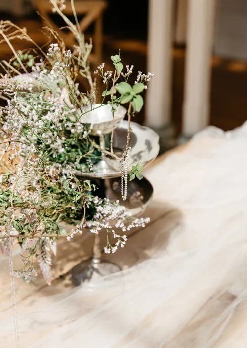 A delicate flower arrangement with white and green flowers and foliage displayed on a tiered silver stand. The display sits on a soft, white fabric background with a pair of dangling silver earrings hanging from the top tier.