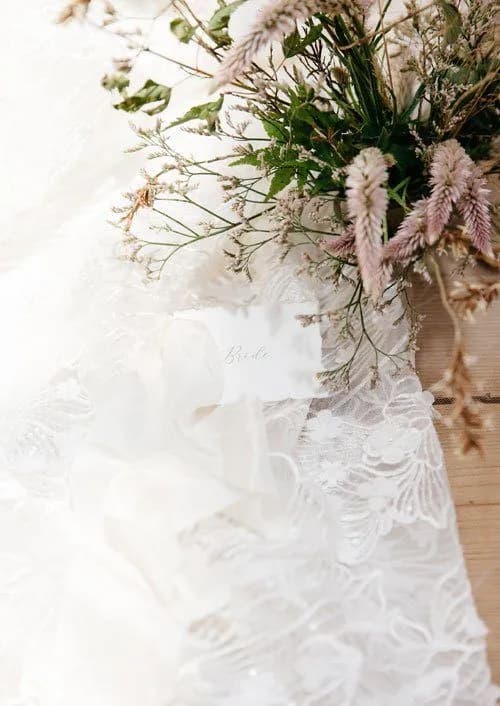 A delicate arrangement of dried flowers lies atop a surface adorned with white lace fabric. A small card labeled "Bride" is nestled among the lace and flowers. The scene exudes a soft, romantic ambiance.