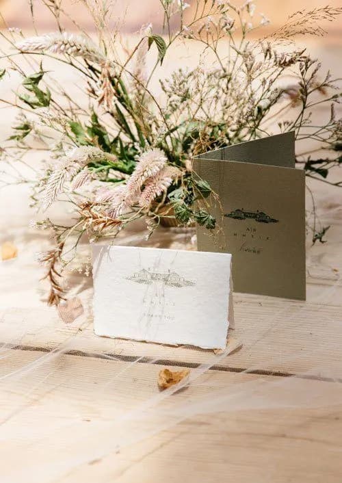 A wedding invitation suite is displayed on a light wooden surface with delicate, dried floral arrangements in the background. The suite includes a textured white card and an olive green folded card, both adorned with elegant, minimalistic designs.