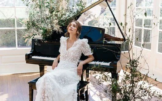 A bride in a white lace wedding dress sits on a stool with her back leaning against a grand piano. She is in a bright room with large windows and surrounded by lush greenery and floral arrangements.