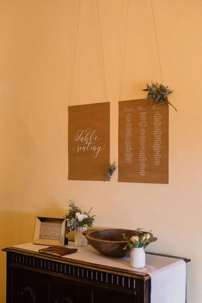 A brown wooden dresser adorned with a white tablecloth features a decorative bowl, small floral arrangements, and a framed picture. Above it, two hanging brown paper scrolls display table seating arrangements, bordered with small bouquets of green foliage.