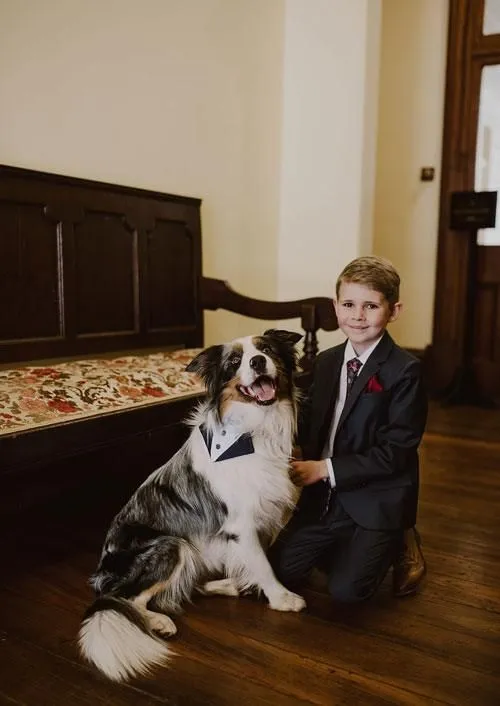 A young boy in a suit with a red boutonniere kneels next to a large dog wearing a bow tie. Both are indoors on a wooden floor, with a vintage wooden bench and a rug in the background. The boy has his arm around the dog, and they are both looking at the camera.