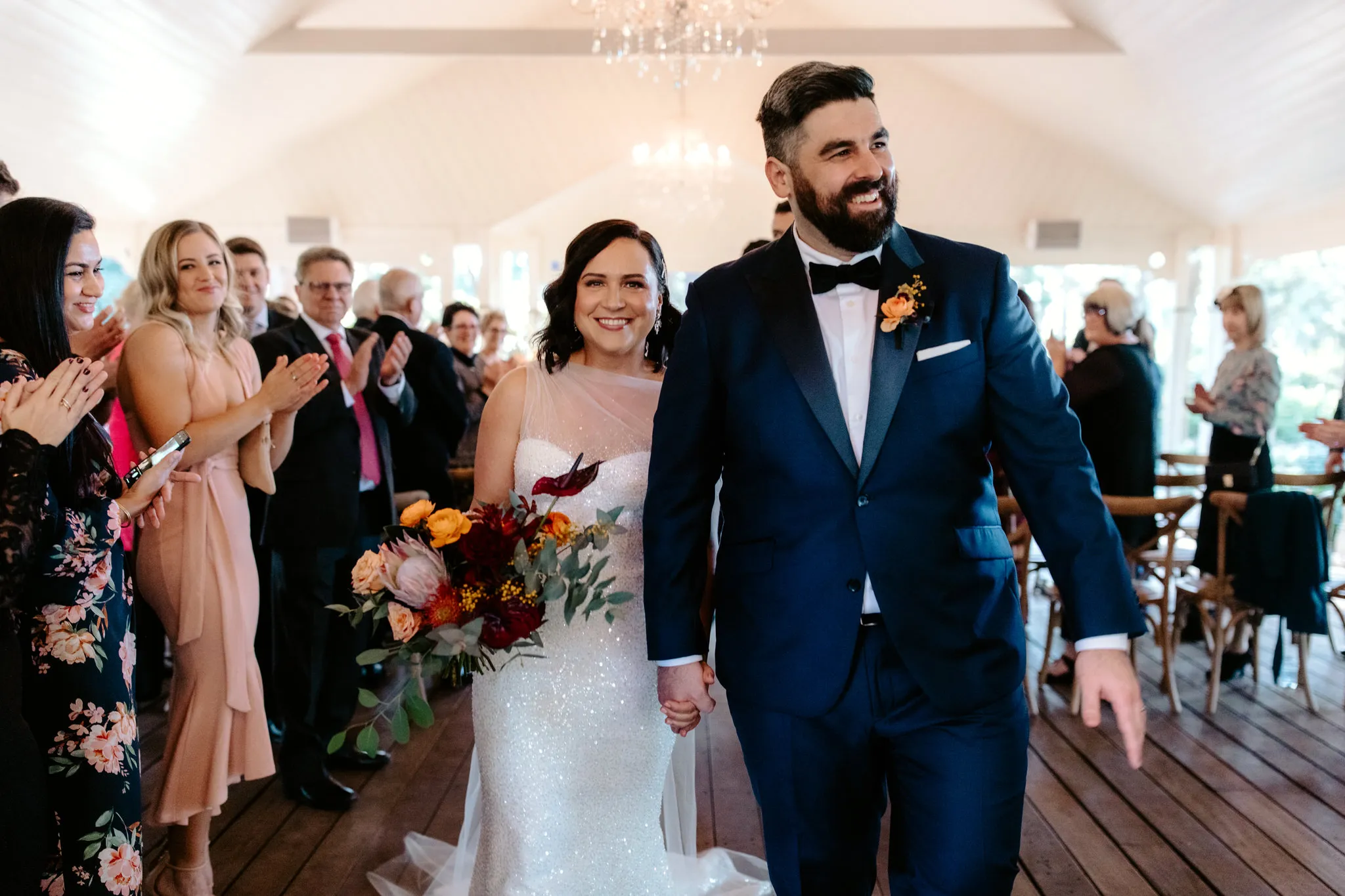A bride and groom walk down the aisle holding hands and smiling. The bride is in a shimmering white gown holding a bouquet of flowers, and the groom is in a navy blue suit with a bow tie. Guests stand and applaud on either side in a well-lit, elegant venue.