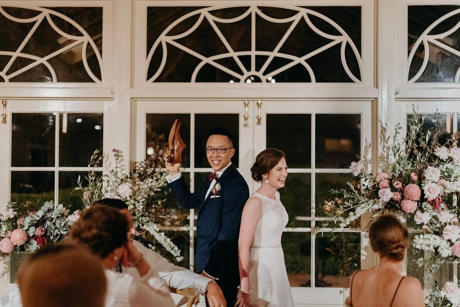 A bride and groom stand back-to-back during a wedding reception game, each holding a shoe in the air. They are in a well-decorated room with large windows and floral arrangements. Guests are seated around them, watching and smiling.