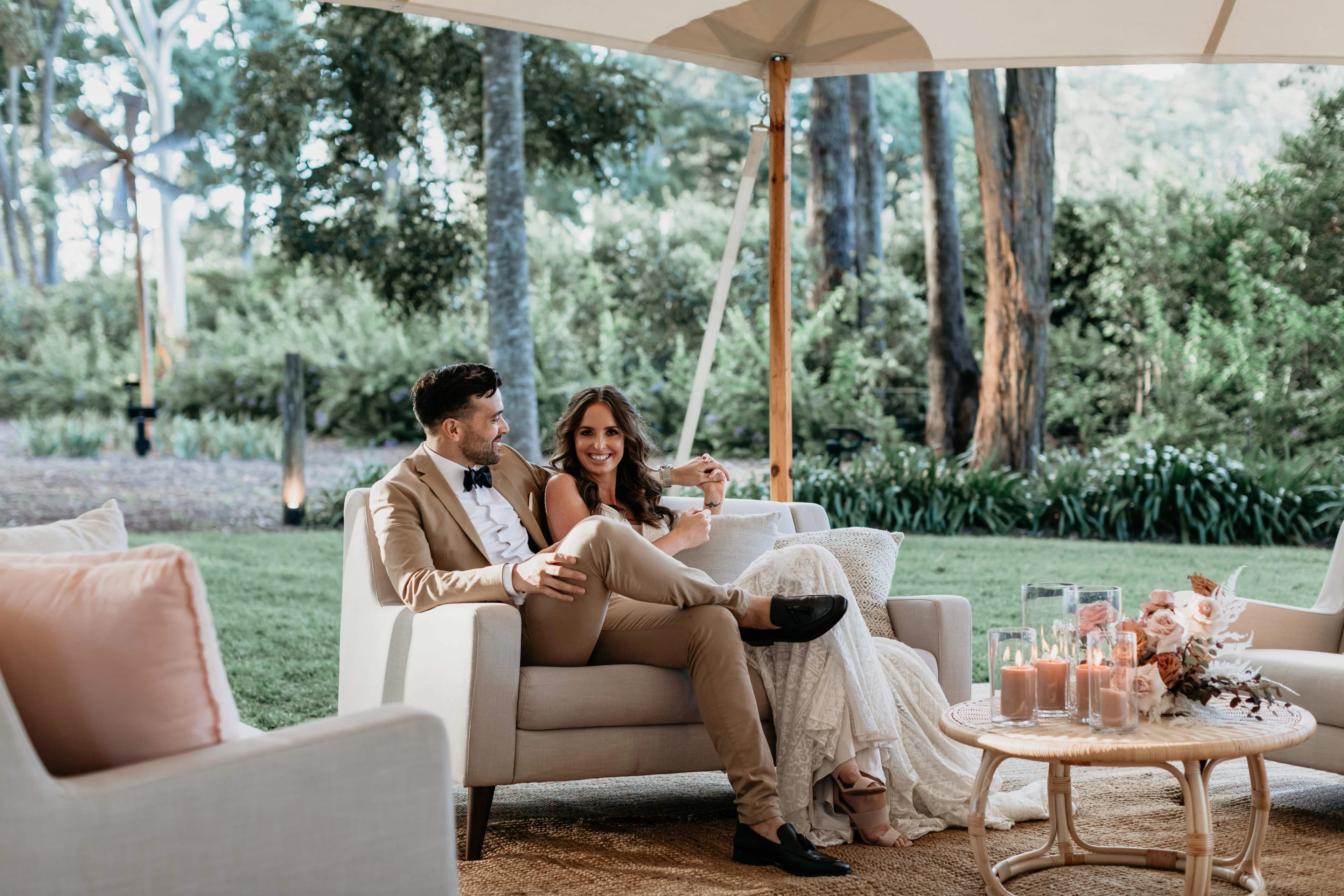 A couple sits on a cushioned outdoor sofa under a canopy in a lush garden setting, both dressed in formal attire. The woman holds a drink and smiles at the man, who has his legs crossed. A round table with floral arrangements and candles is in front of them.