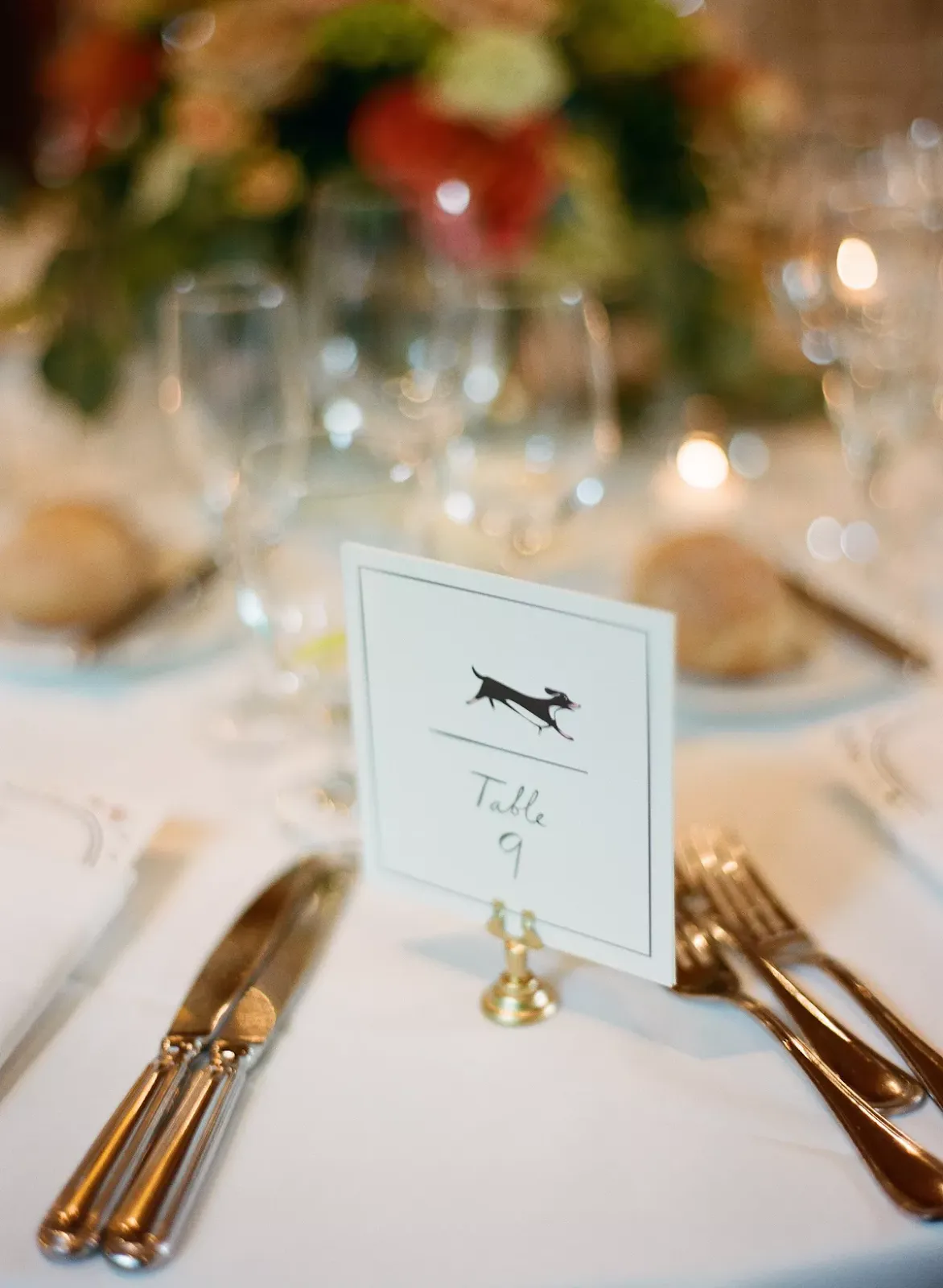 A table setting with a card labeled "Table 9" held by a small gold stand featuring a dog illustration. The table is set with silverware, wine glasses, and bread rolls, and adorned with a blurred background of greenery and flowers.