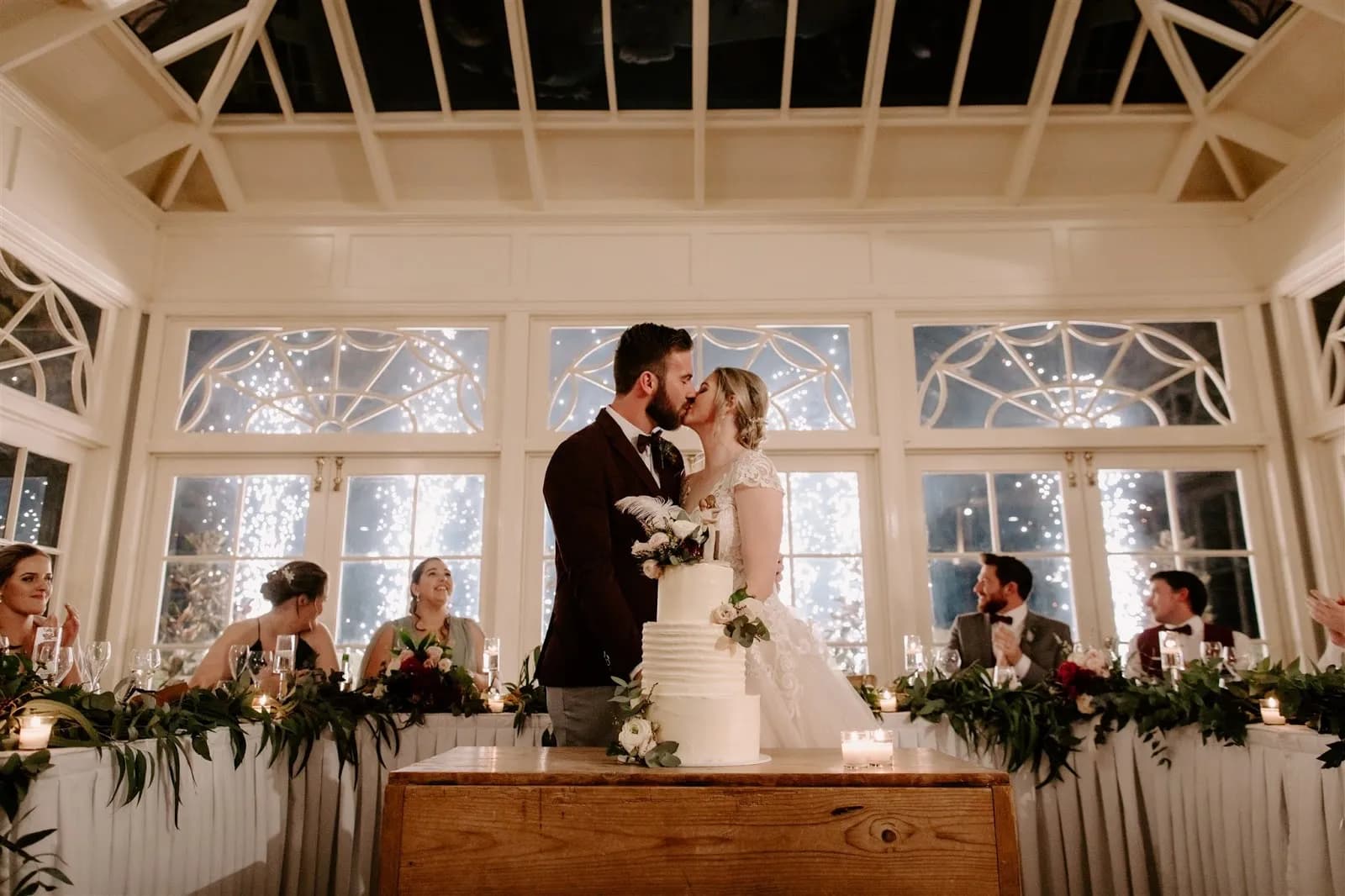 A bride and groom share a kiss behind their wedding cake in a beautifully decorated room with a glass ceiling. Bridesmaids and groomsmen are seated at a table covered with greenery and candles, and twinkling lights illuminate the background.