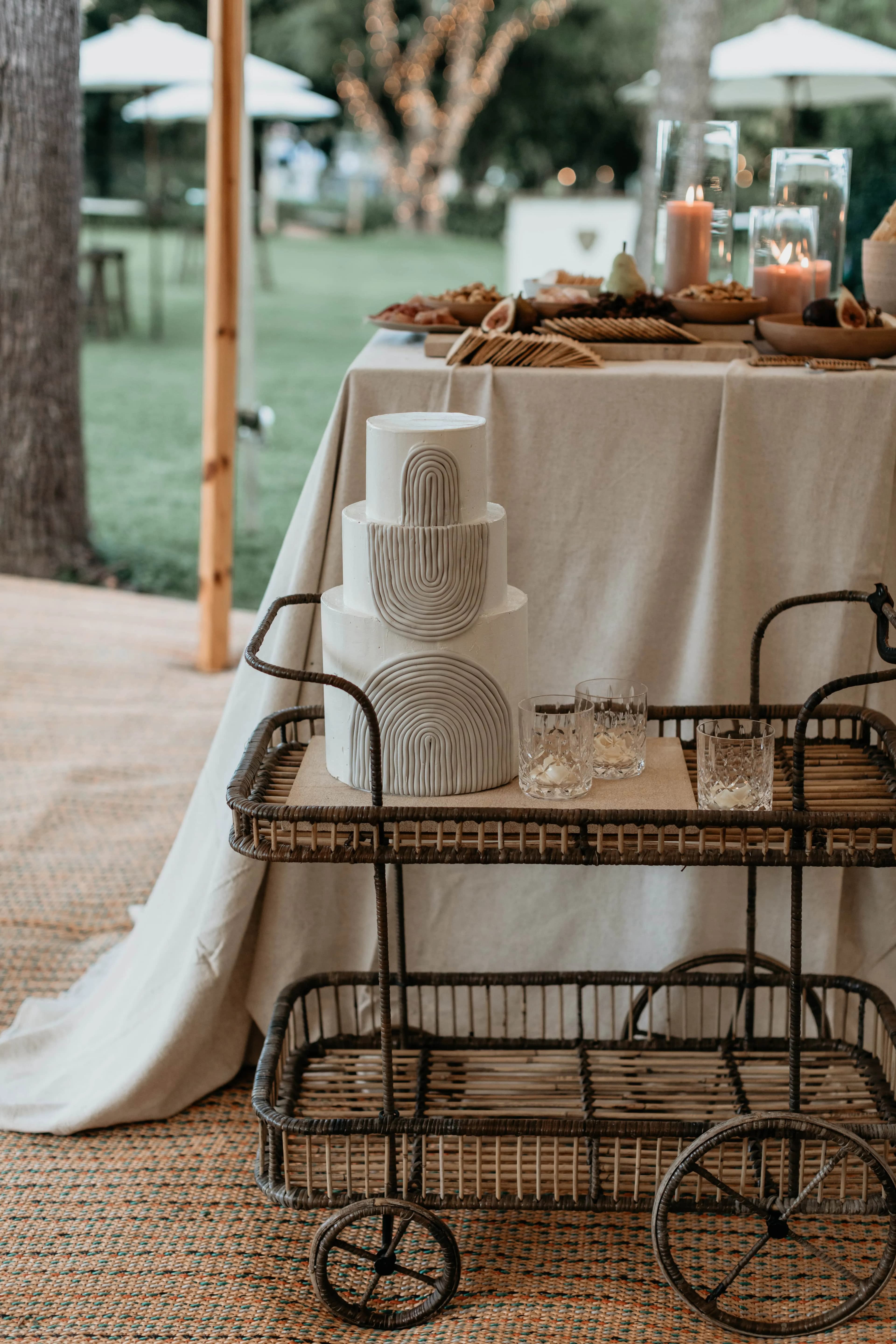 A two-tier white wedding cake with a modern geometric design sits on a wicker cart with glassware. The cart is in front of a table covered in a white tablecloth adorned with candles and food, set outdoors with blurred greenery and patio umbrellas in the background.