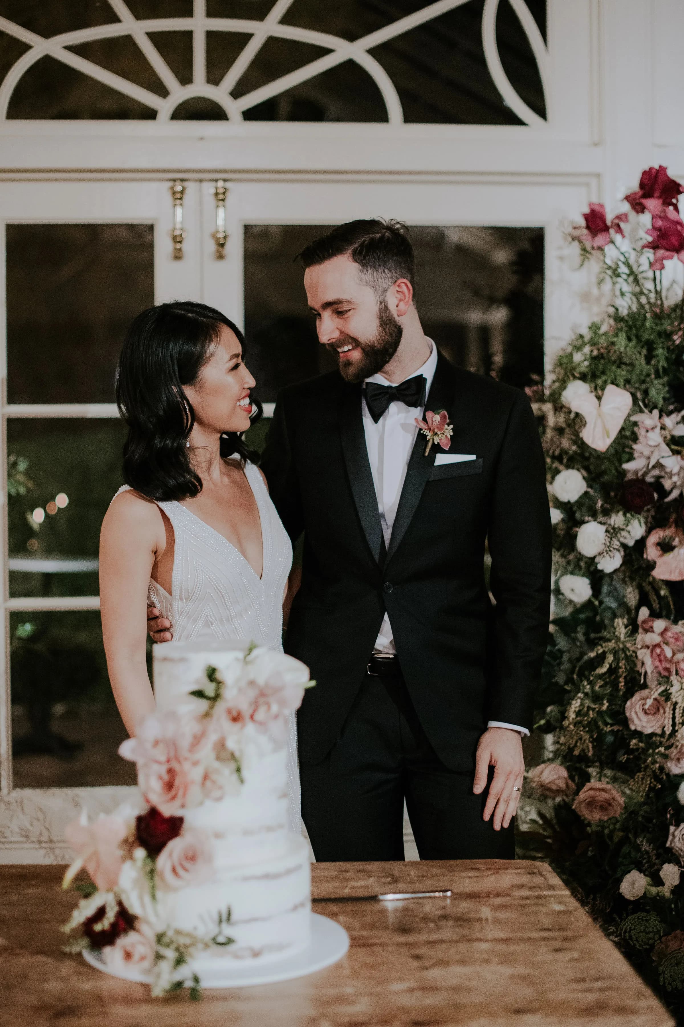 A couple stands together, smiling at each other in front of a decorated white table with a wedding cake. The woman is wearing a white dress, and the man is in a suit with a bow tie. Behind them, there is greenery and white flowers, framed by a window.